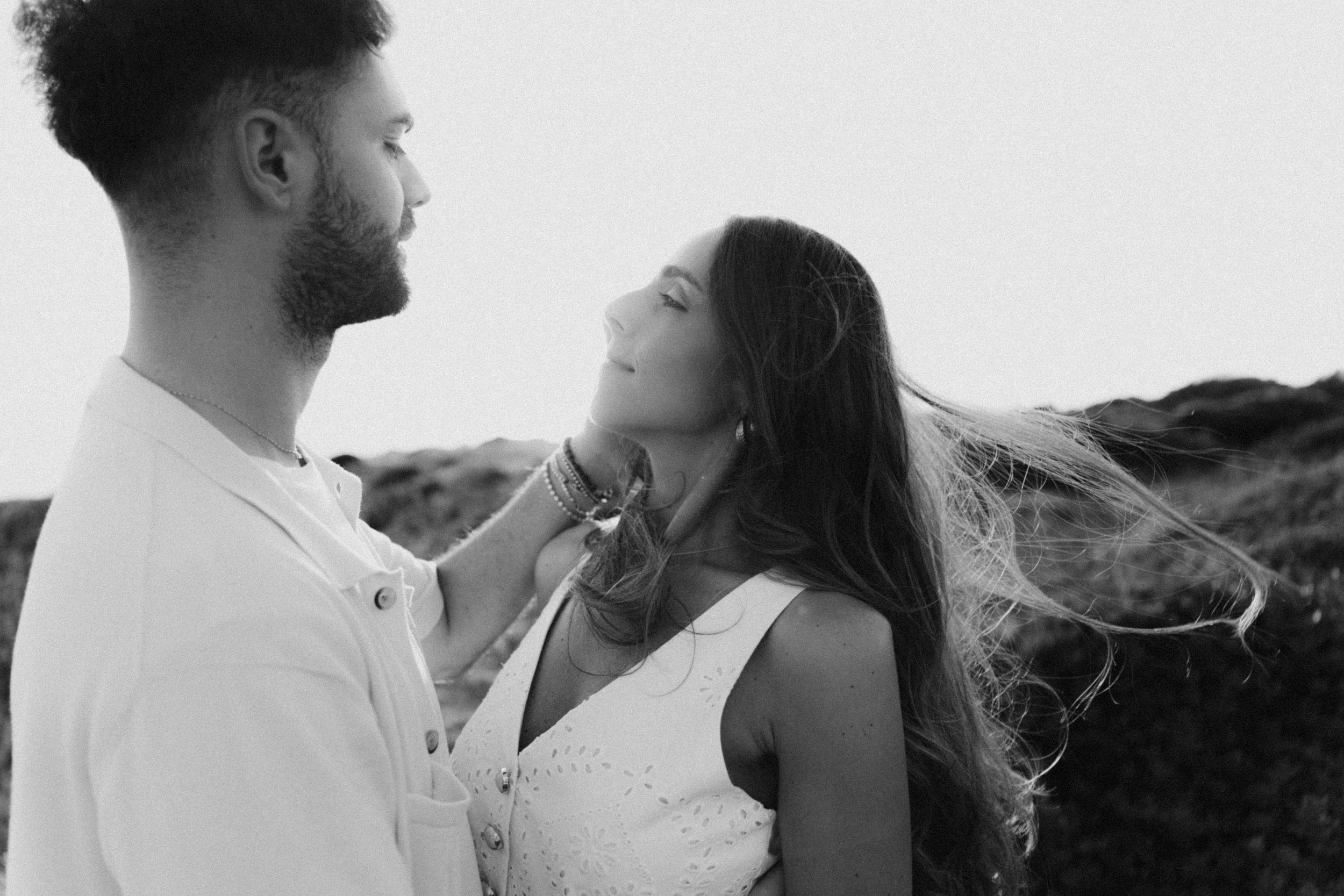 Black and white photo of a couple sharing a tender moment outdoors, woman's hair blowing in the wind.