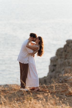 A couple sharing a romantic kiss on a coastal cliff at sunset, with the ocean shimmering in the background.