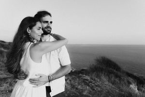 Black and white photo of a couple embracing on a coastal cliff with ocean views in the background.
