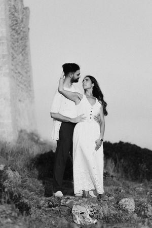Black and white photo of a couple embracing near ancient ruins, woman in white dress gazing up at man.