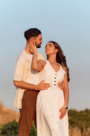 Couple shares tender moment at sunset, woman in white dress gently touches mans face outdoors.