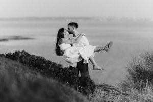 Man lifting woman for a kiss outdoors near a lake in a romantic black and white engagement photo.