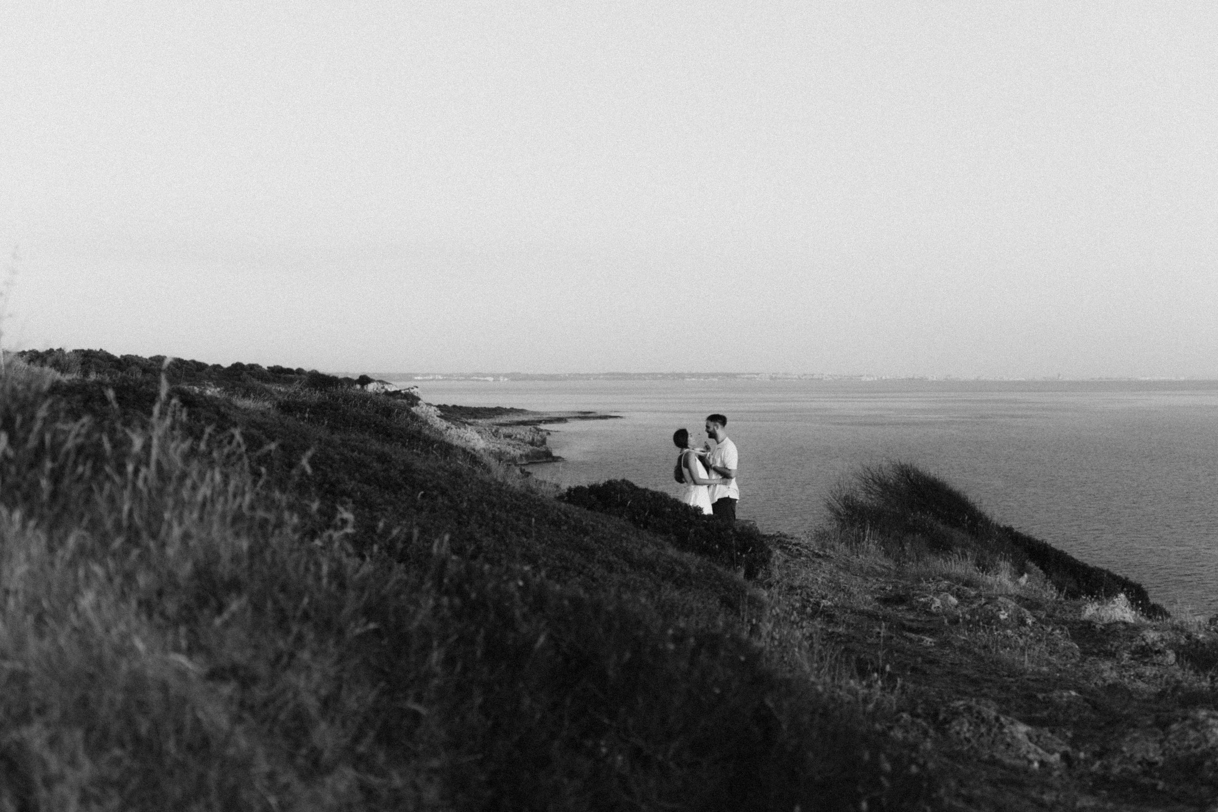 Black and white image of a couple embracing on a grassy coastal hillside overlooking the sea.