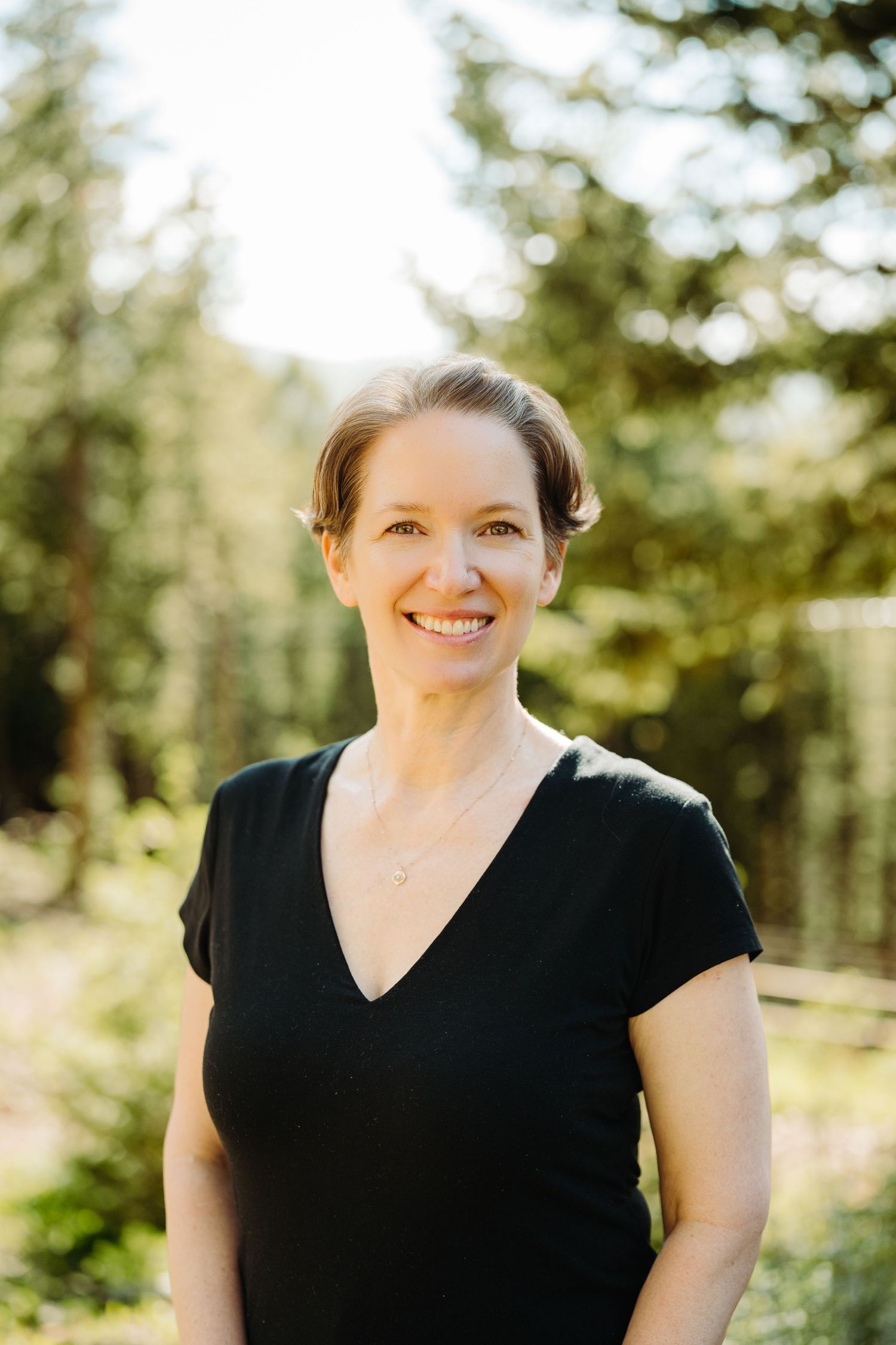 Smiling woman in black v-neck top standing outdoors among tall pine trees in natural sunlight by McGlone Photography