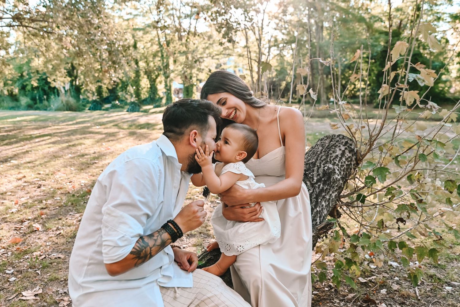 A young family with tattoed father, mother in white dress, and baby enjoying a sunny outdoor photo session in a lush park.