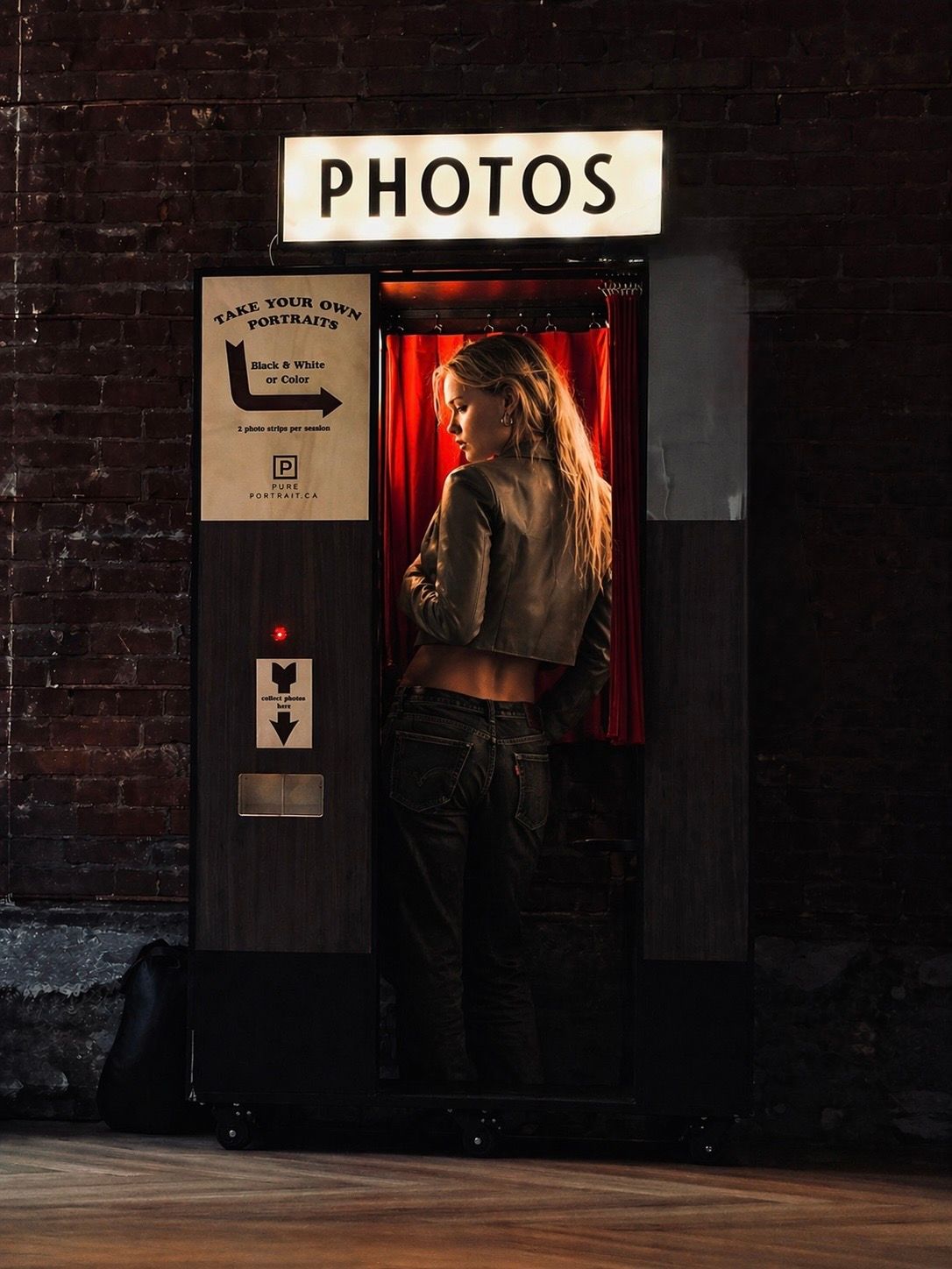 Woman posing inside a vintage photo booth with red curtain and glowing PHOTOS sign against brick wall.