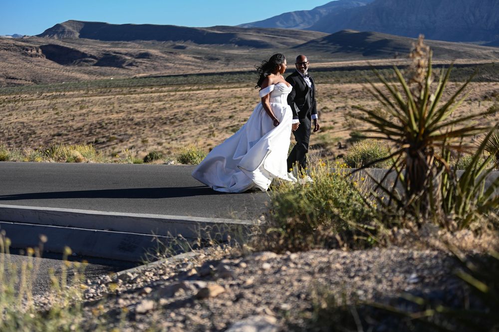 Bride and groom walk along desert road in wedding attire with mountains and yucca plants in background.