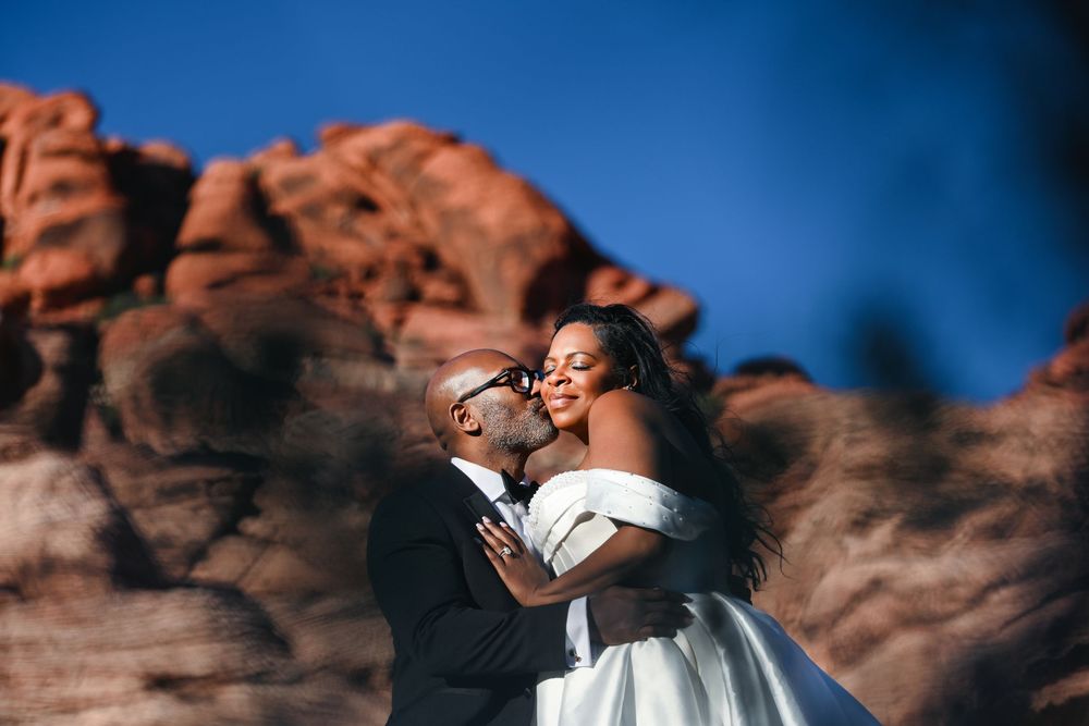 Couple shares romantic kiss at Red Rock Canyon wedding, bride in off-shoulder gown, groom in black suit, desert backdrop.