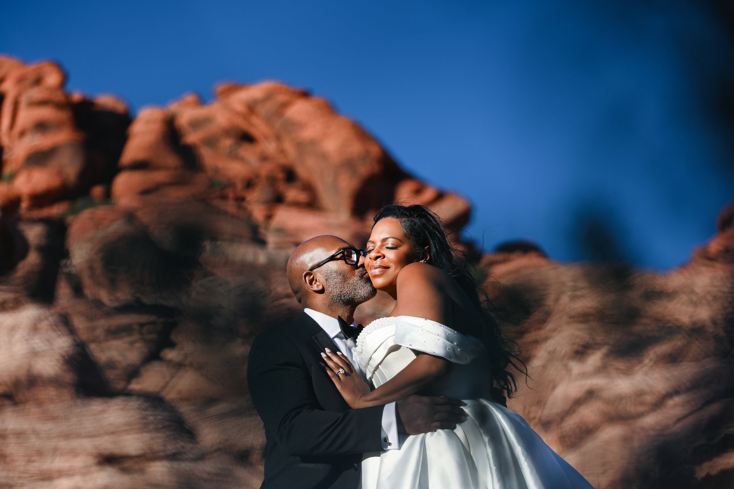 Couple shares romantic kiss at Red Rock Canyon wedding, bride in off-shoulder gown, groom in black suit, desert backdrop.