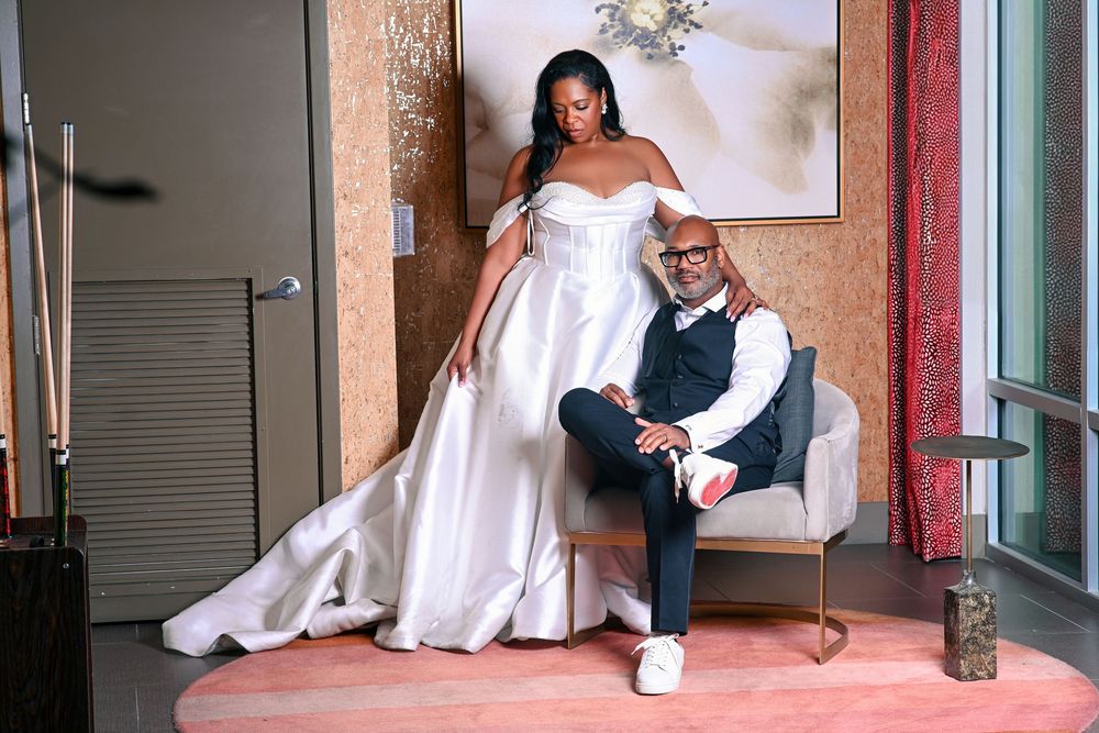 Bride in white off-shoulder gown stands beside groom seated in chair, elegant wedding portrait indoors.