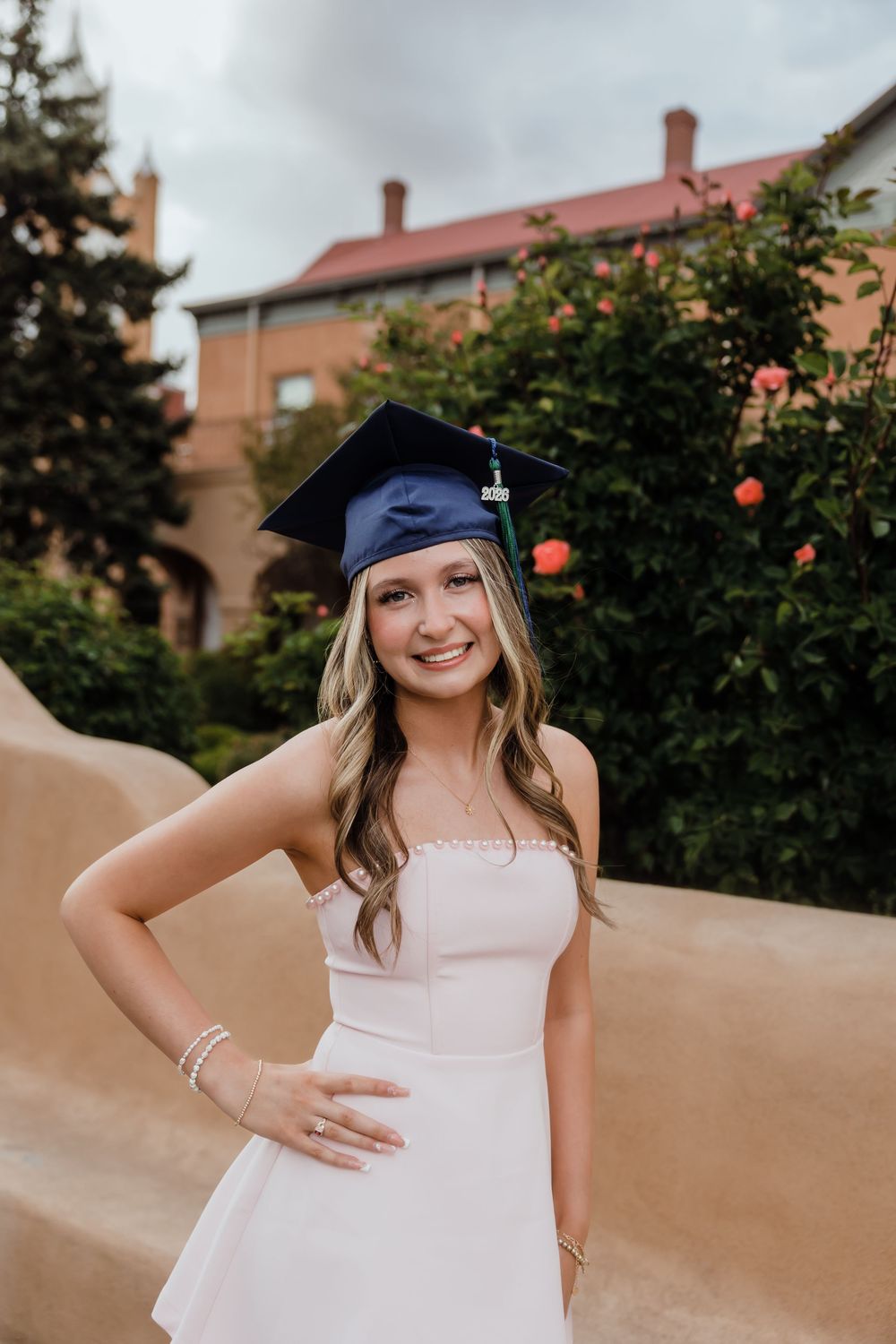 Young woman in white dress and blue graduation cap smiling outdoors near flowering bushes and brick building.