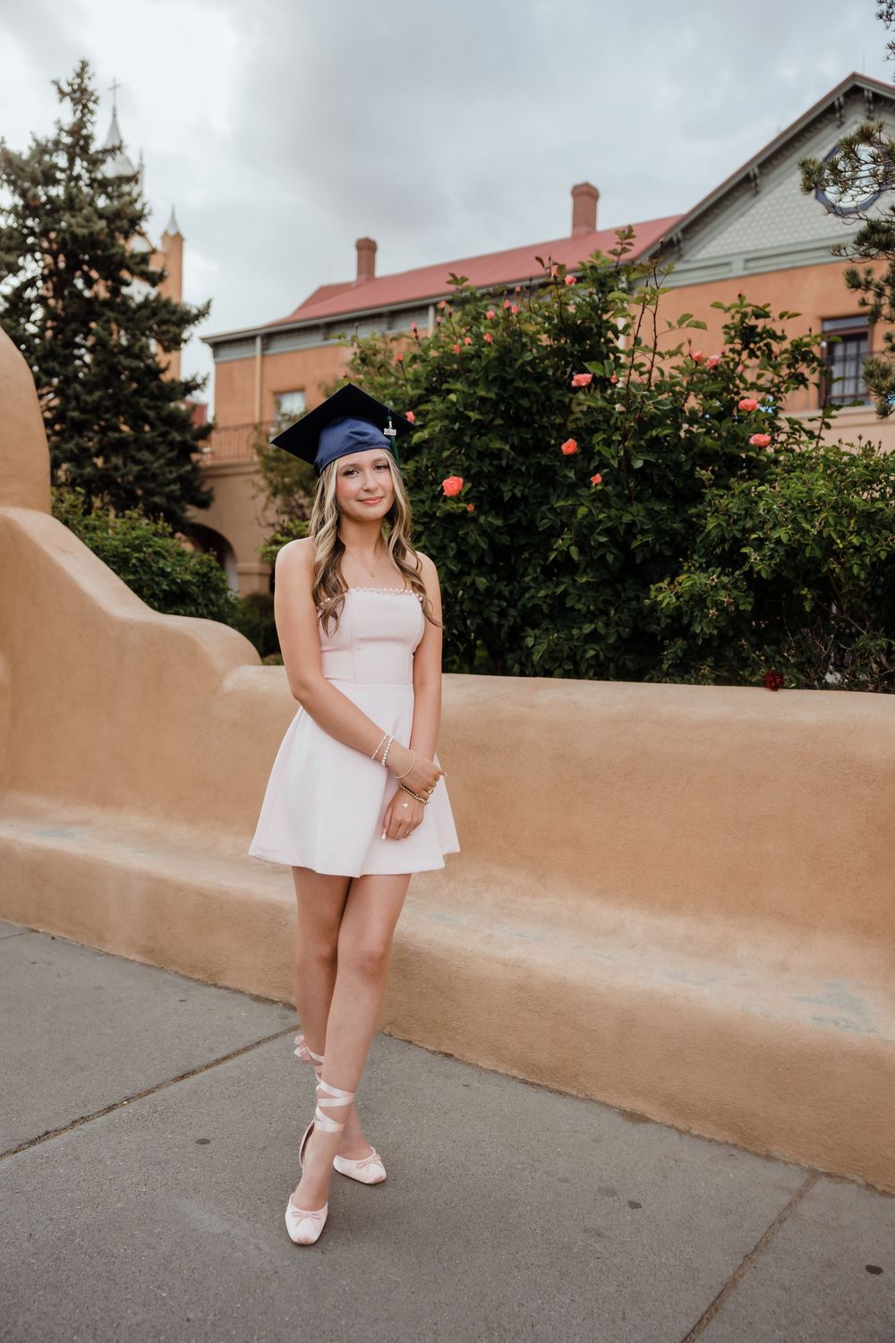 Young woman in white dress and blue graduation cap poses outdoors near adobe wall with roses.