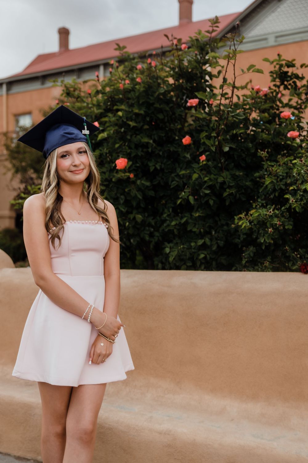 Smiling graduate in white dress and blue cap stands before blooming rose bushes and brick building.