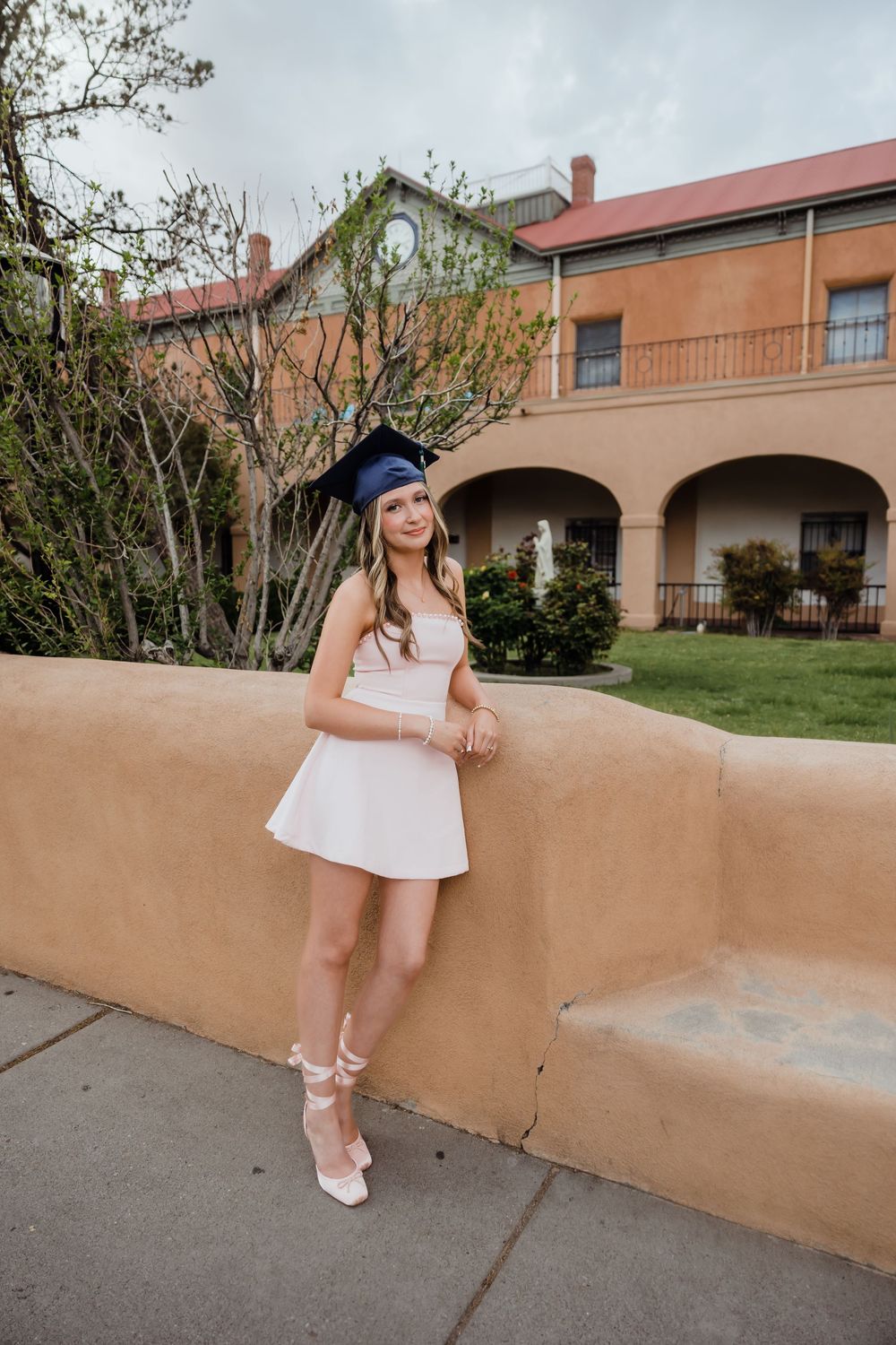 Young woman in white dress and graduation cap poses by adobe wall at university campus.