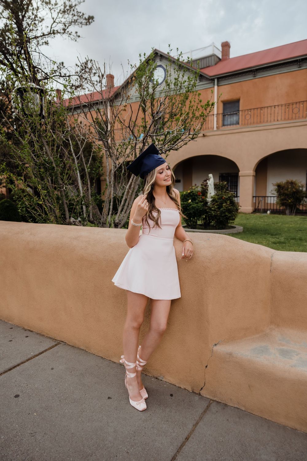 Young woman in white mini dress and heels holds graduation cap outdoors near adobe building.