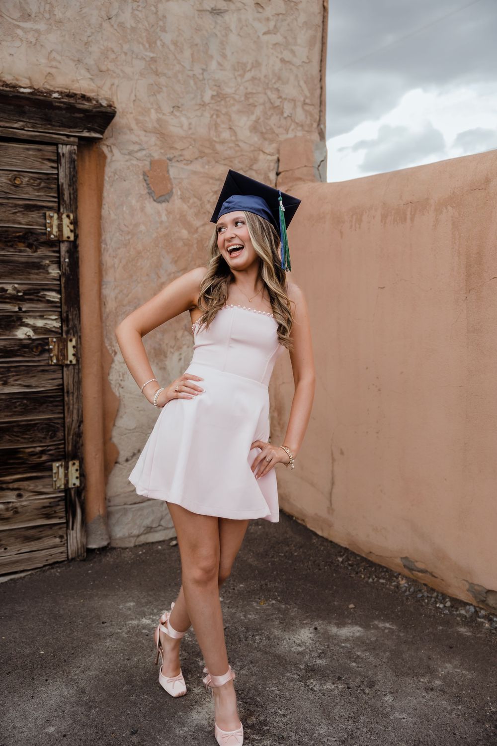 Young woman in white dress and blue graduation cap poses confidently outdoors near adobe wall.