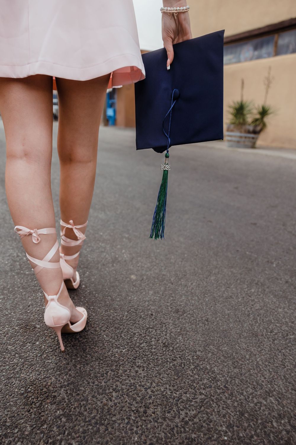 Close-up of graduate walking in pink heels holding navy blue graduation cap with green tassel.