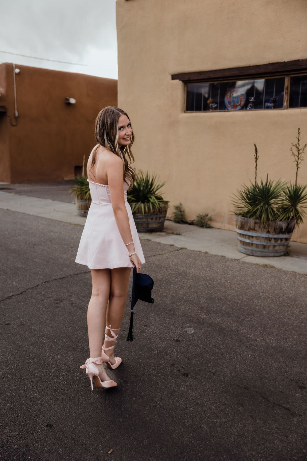 Graduate in white dress carries blue cap while walking past adobe building with potted plants.