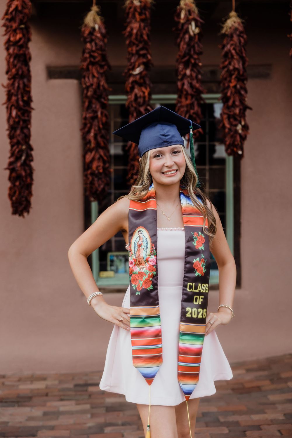 Smiling graduate in navy cap and colorful Class of 2026 stole poses in front of hanging red chile ristras.