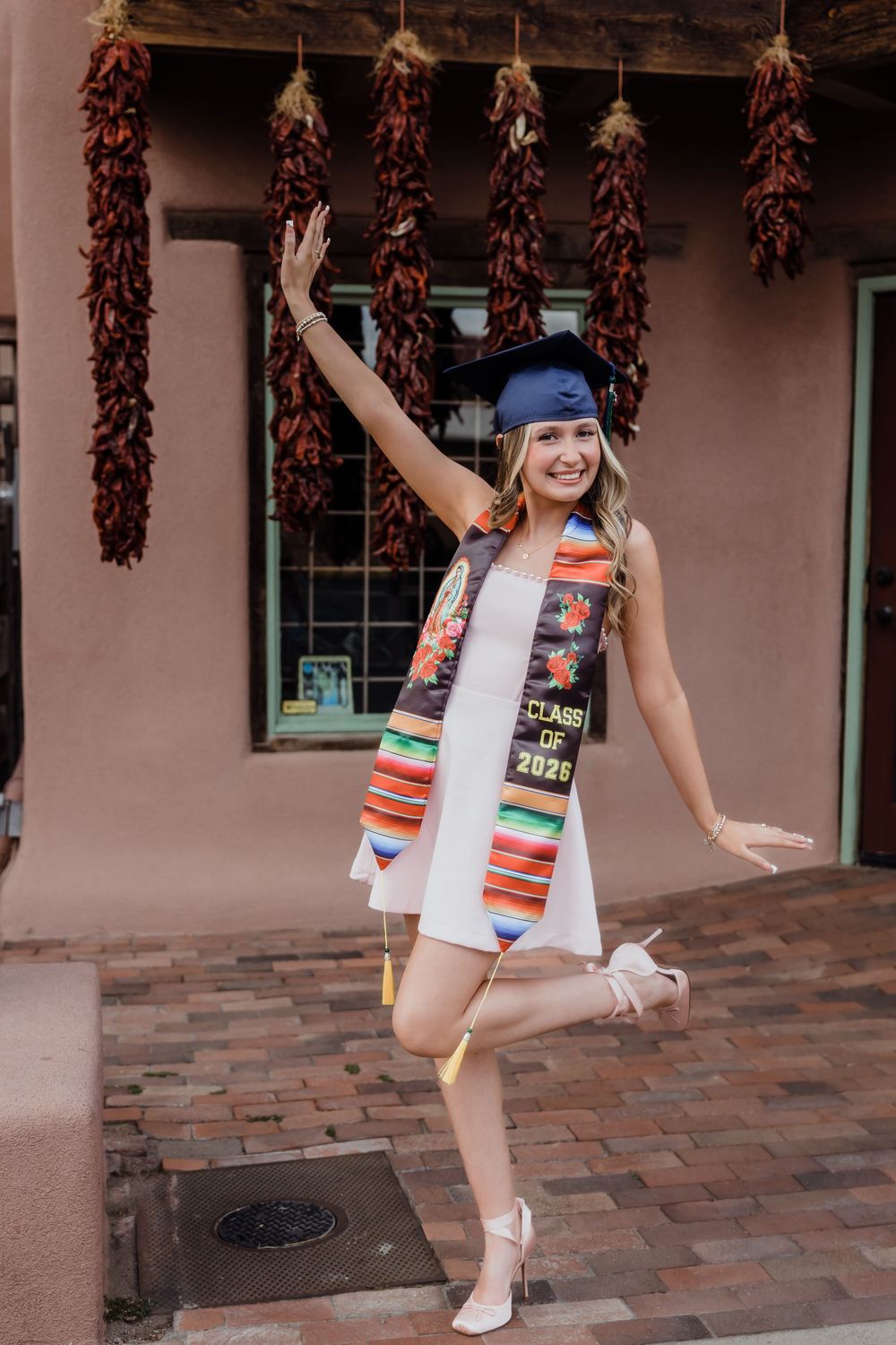 Smiling graduate in white dress and blue cap celebrates with colorful stole reading Class of 2025.
