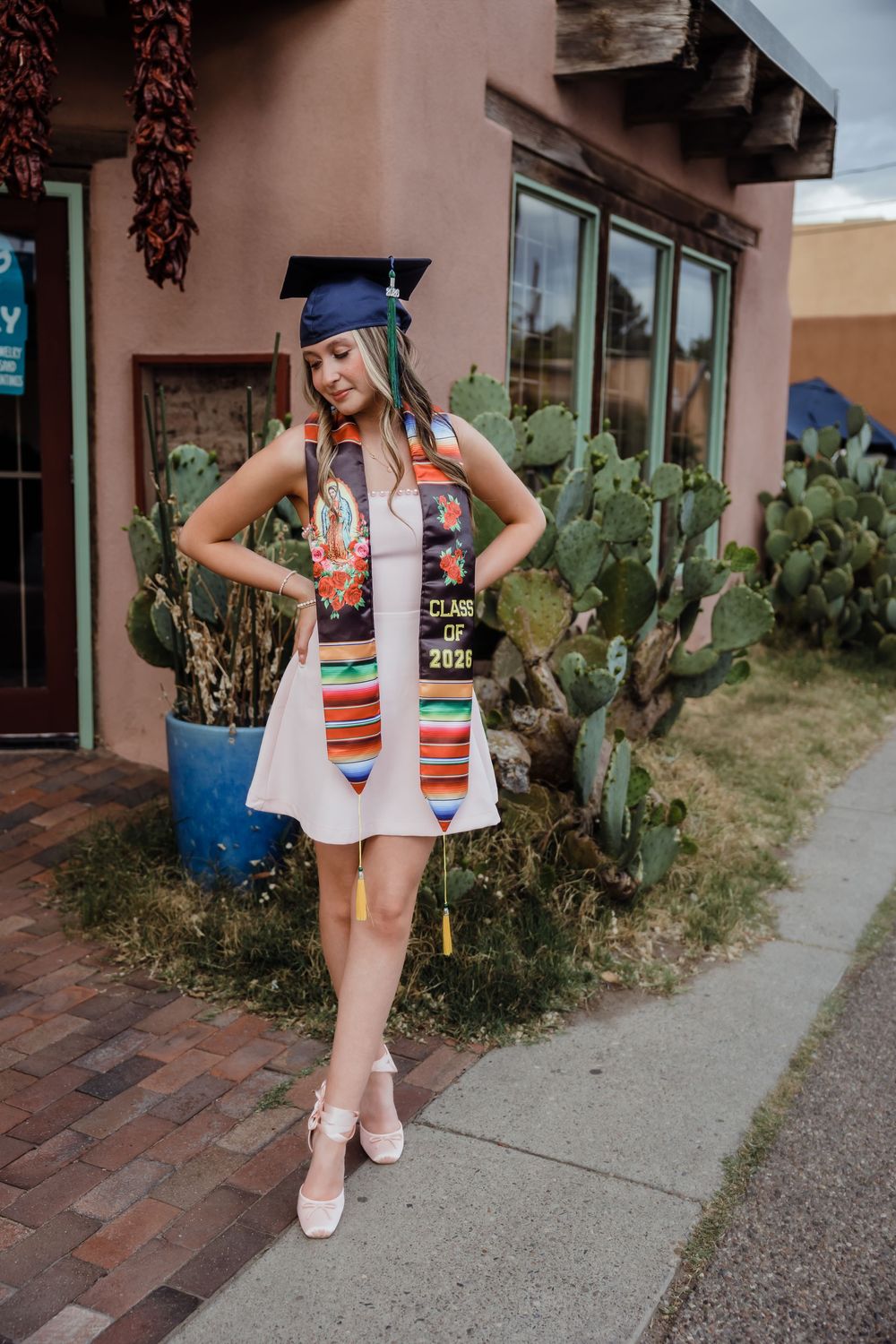 Graduate in white dress with colorful Class of 2021 stole and navy cap poses by cactus.
