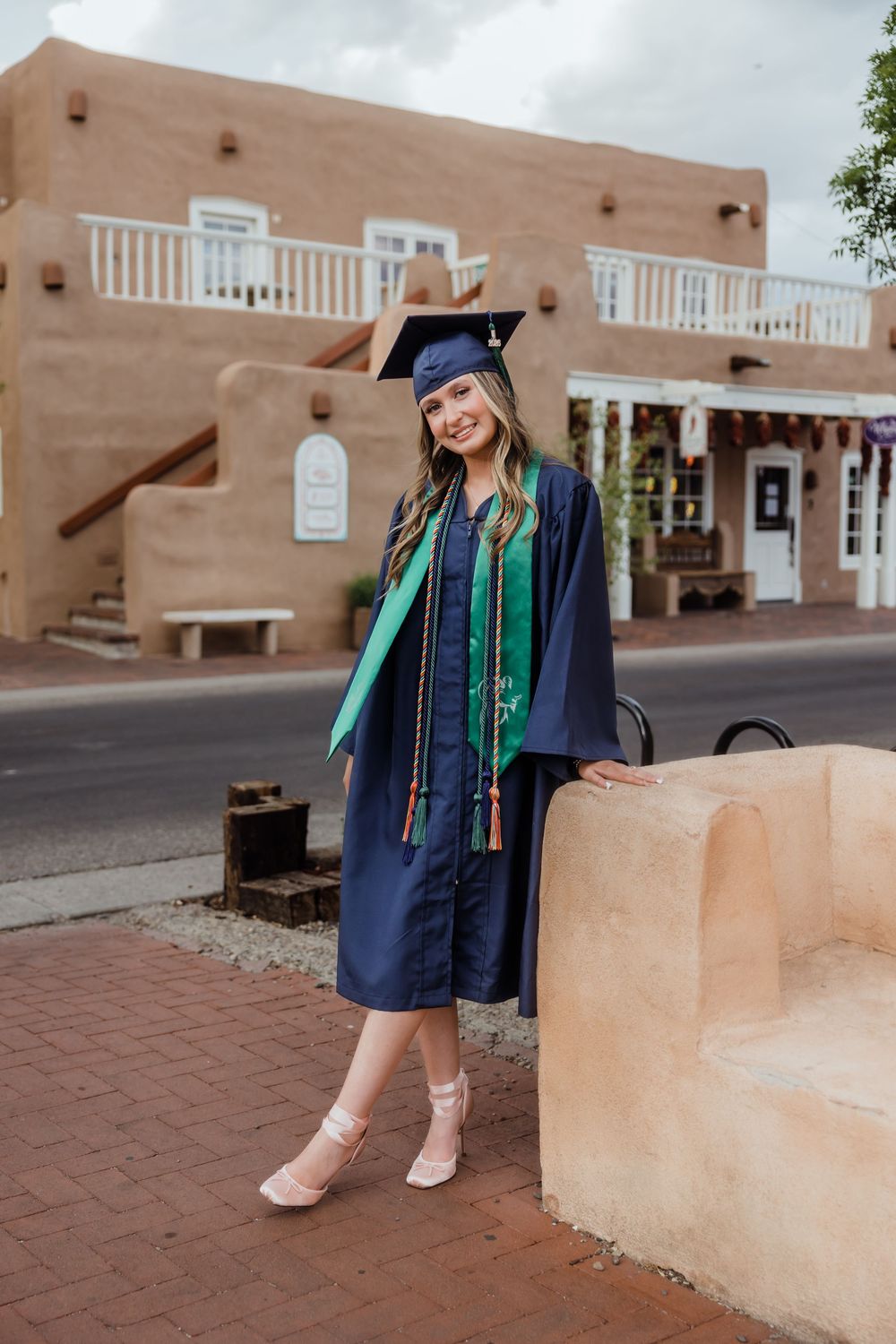 Smiling female graduate in navy cap and gown with green stole posing outdoors in adobe setting.