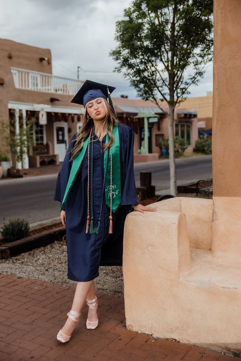 Graduate in navy cap and gown with green stole poses by adobe column on city street.