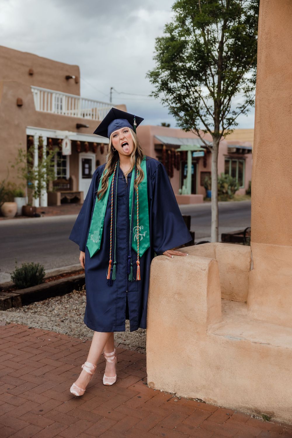 Excited female graduate in navy cap and gown laughing outdoors near adobe building with green stole.
