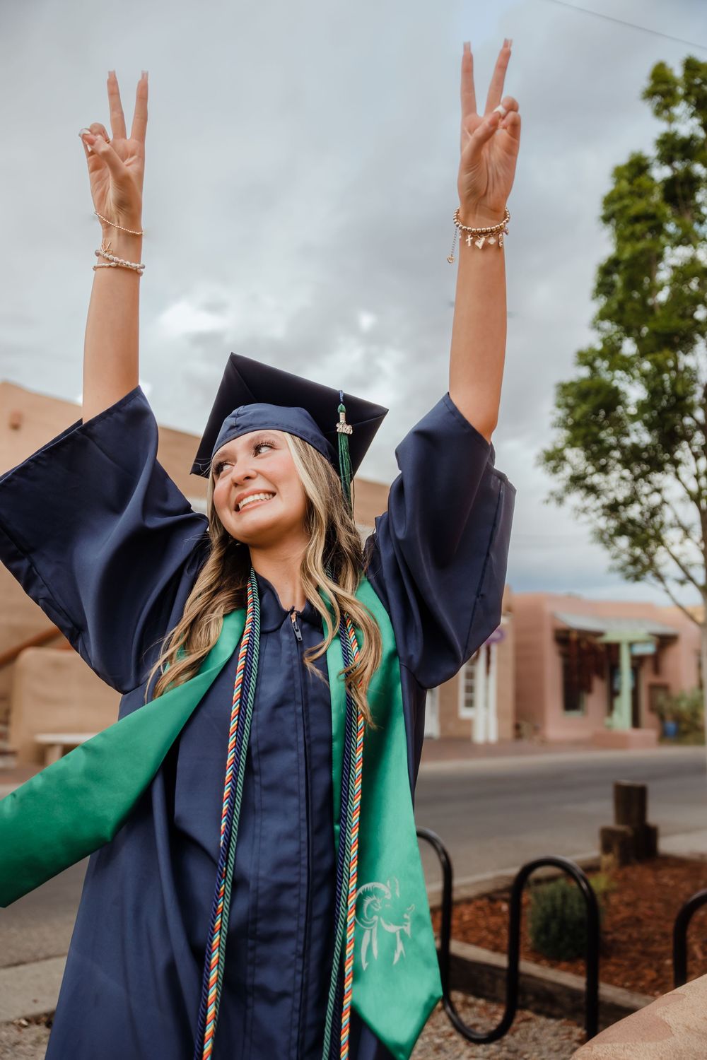 Excited graduate in navy cap and gown raises peace signs with both hands outdoors on cloudy day.