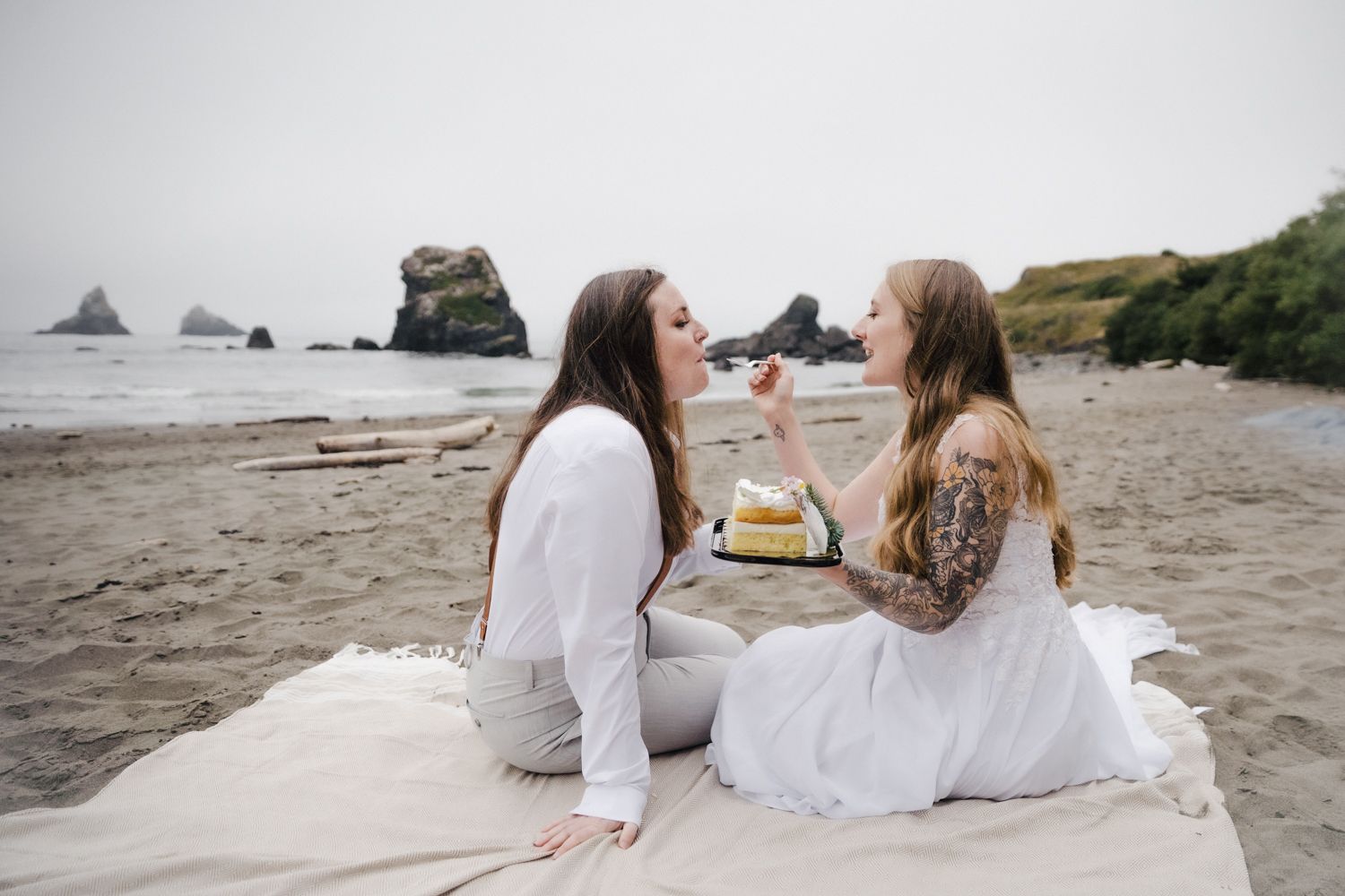 Two brides feed each other wedding cake that they bought from the grocery store and decorated themselves on the beach where they exchanged v