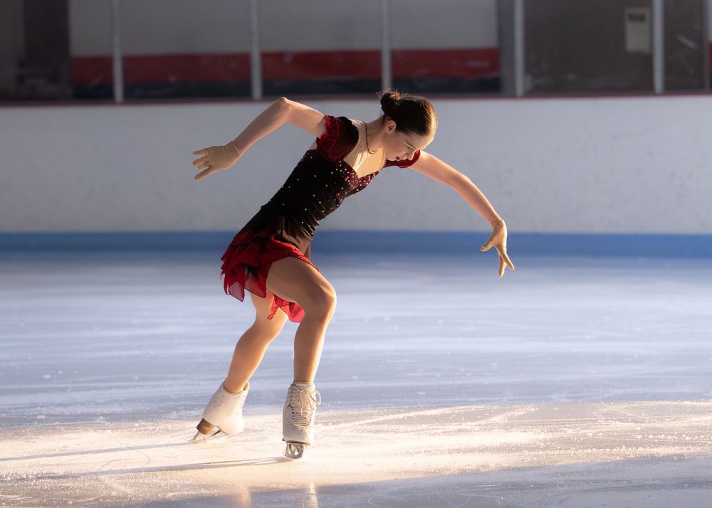 Young female figure skater performing graceful routine in black and red dress on indoor ice rink. Hagerstown, Maryland.