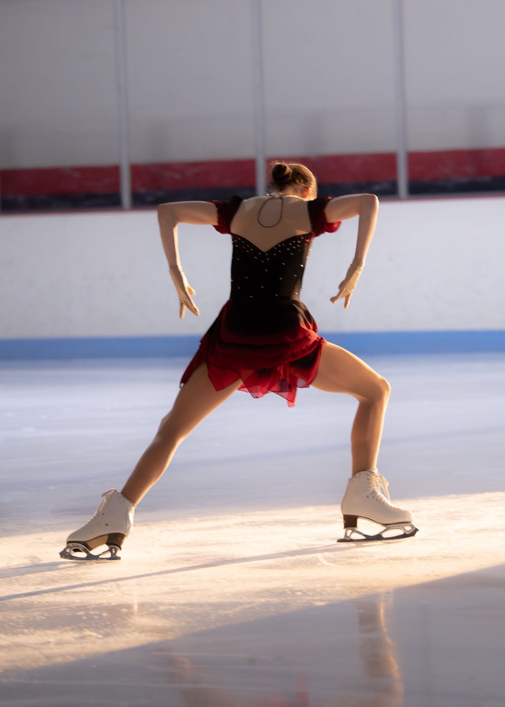 Female figure skater in black and red costume viewed from behind in wide stance on ice rink. Hagerstown, Maryland.