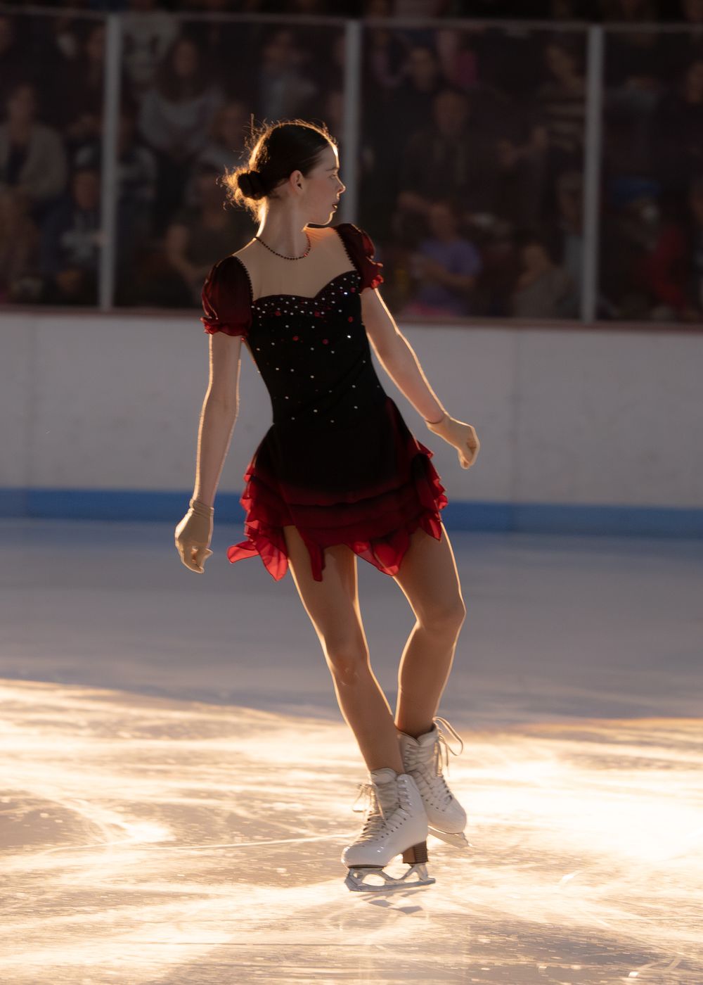 Young female figure skater in elegant black and red dress gliding on ice in evening light.