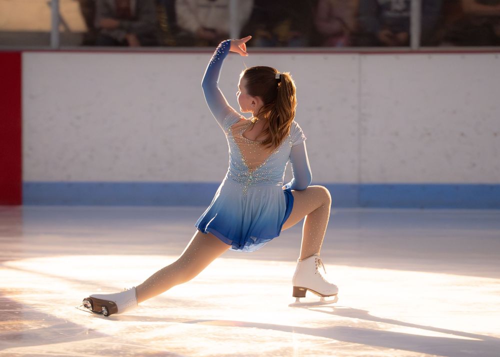 Female figure skater in blue gradient dress performing elegant low split pose on ice rink. Hagerstown, Maryland.