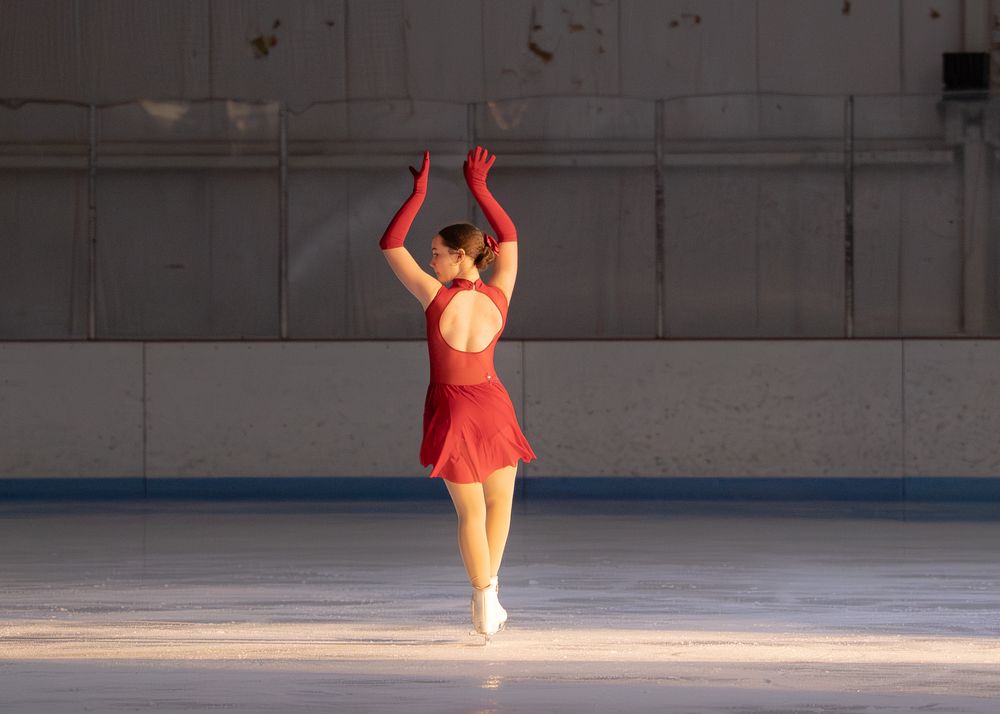 Female figure skater in red dress and gloves performing arabesque spin on outdoor ice rink. Hagerstown, Maryland.