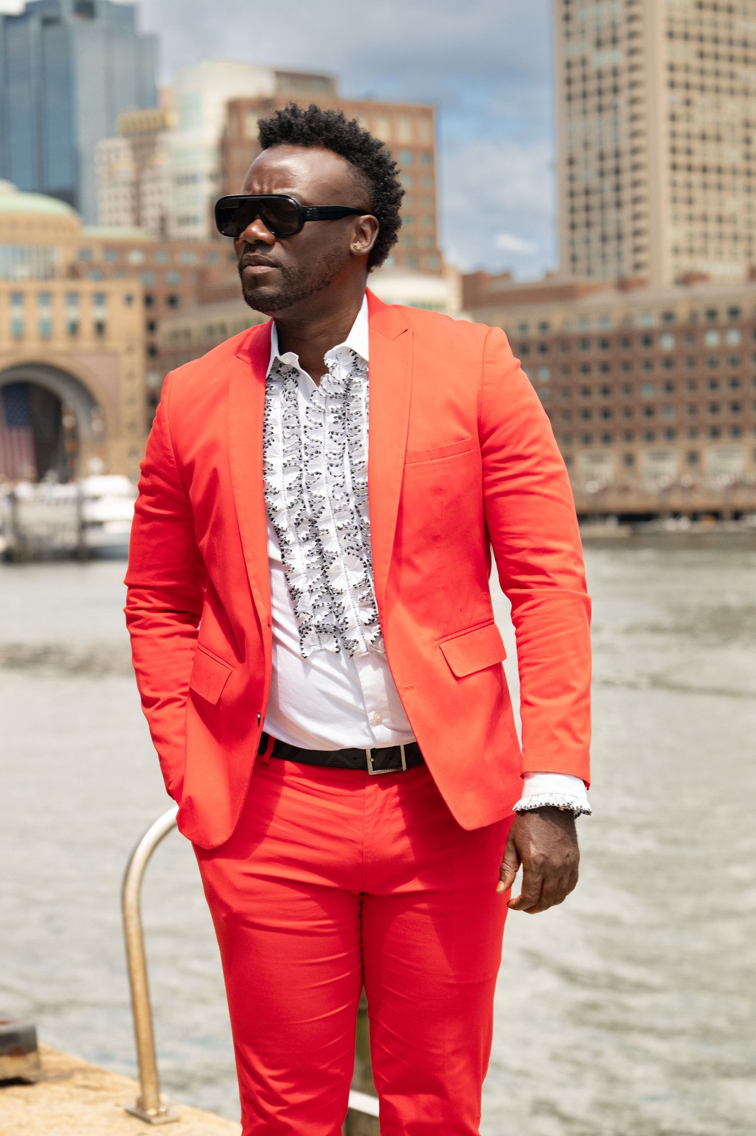 Stylish man in bold red suit and sunglasses posing near waterfront with city buildings in background.