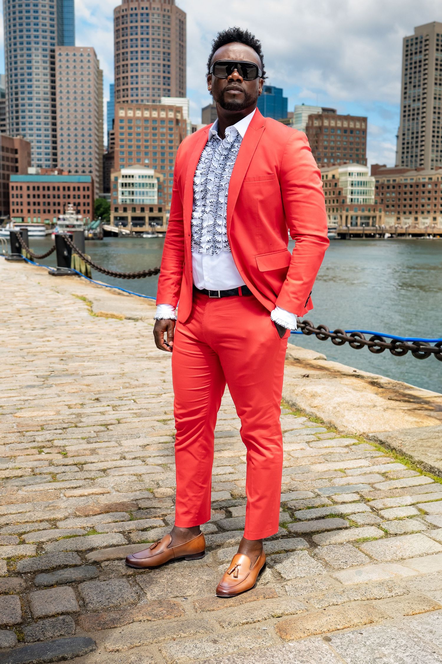 Man in bold red suit with ruffled shirt and tan loafers posing on cobblestone waterfront with Boston skyline.