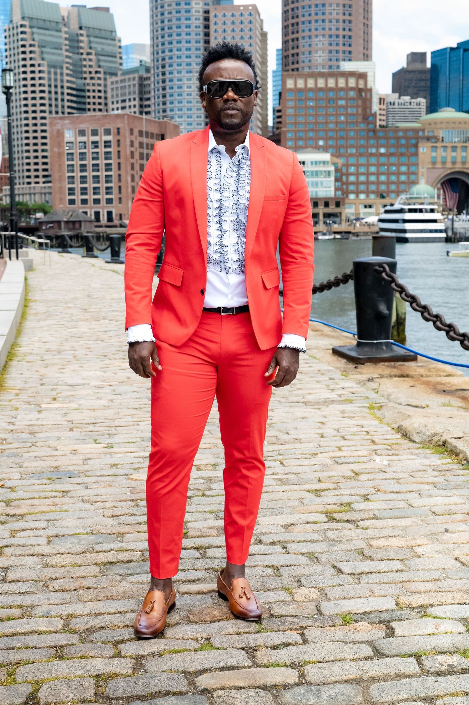 Man in bold red suit and sunglasses standing on cobblestone waterfront with city skyline backdrop.