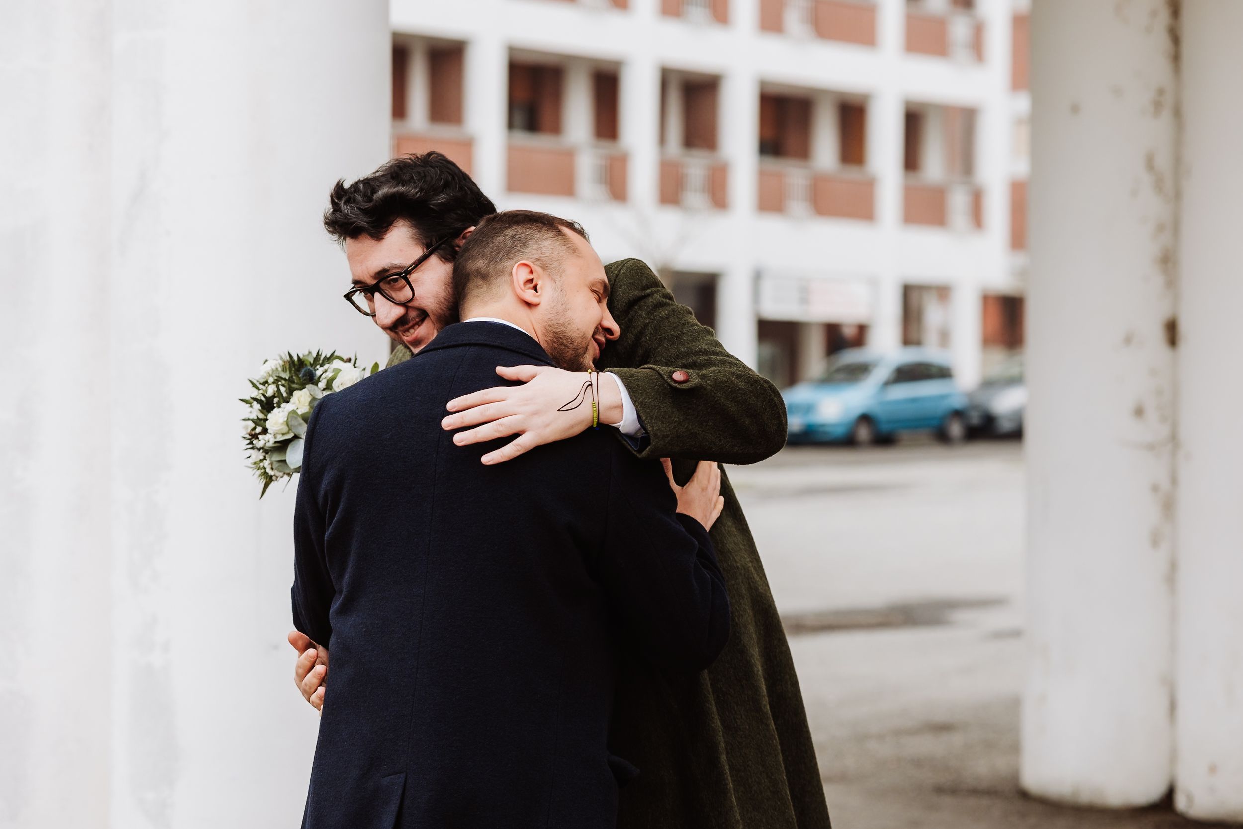 Two men in formal attire share a warm embrace outdoors, one holding a small bouquet, suggesting a wedding celebration.