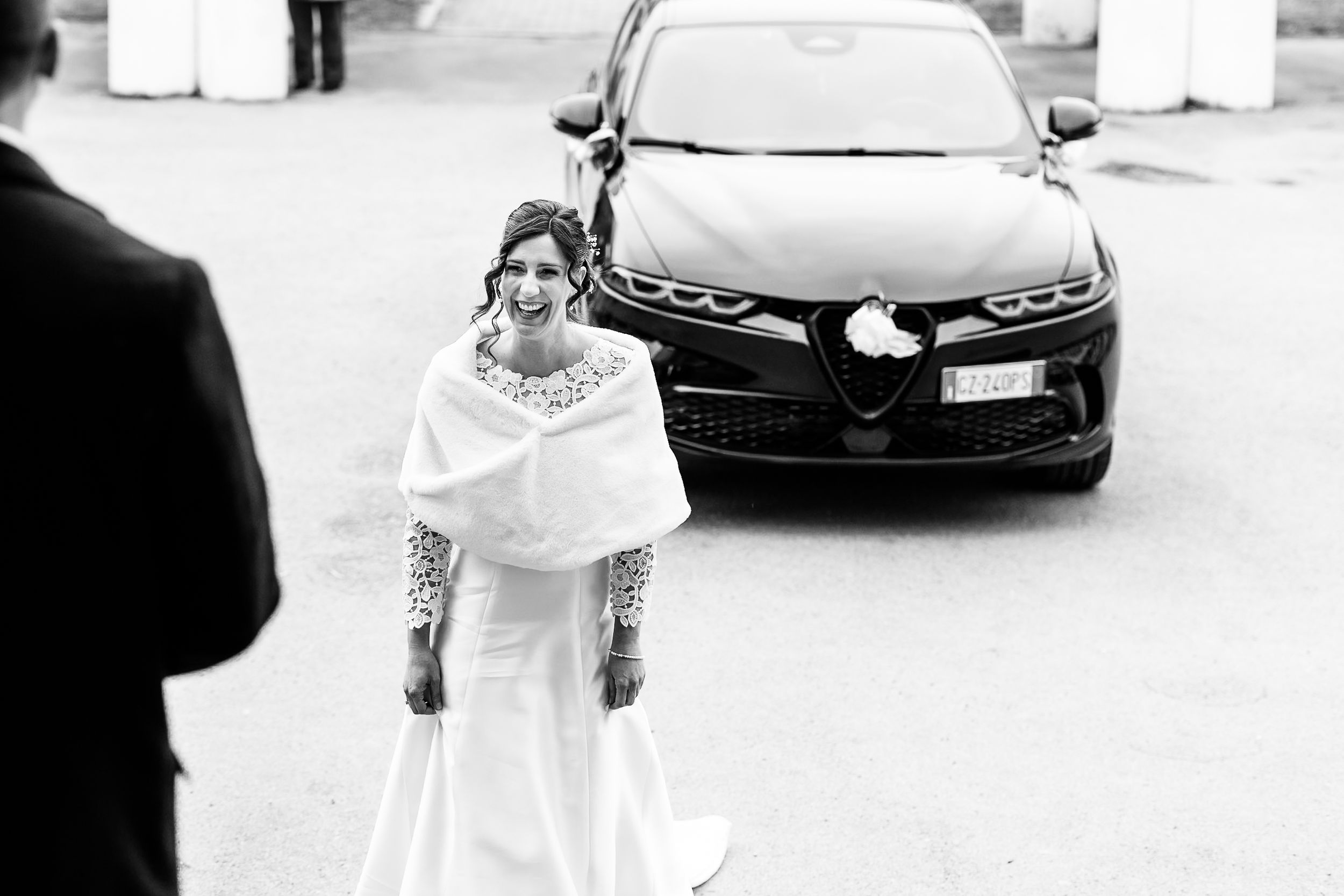 Bride in white gown and cape smiling near black Alfa Romeo car in black and white wedding photo.