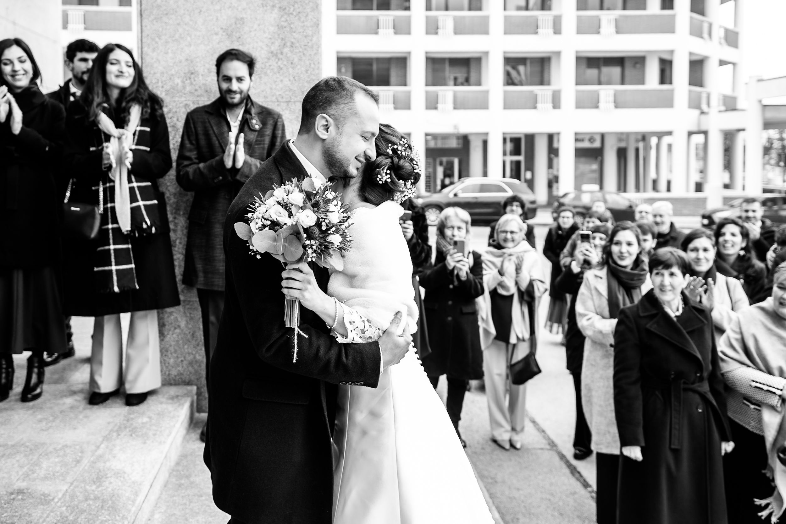 Black and white photo of a newlywed couple sharing a kiss while guests celebrate at an outdoor wedding ceremony.