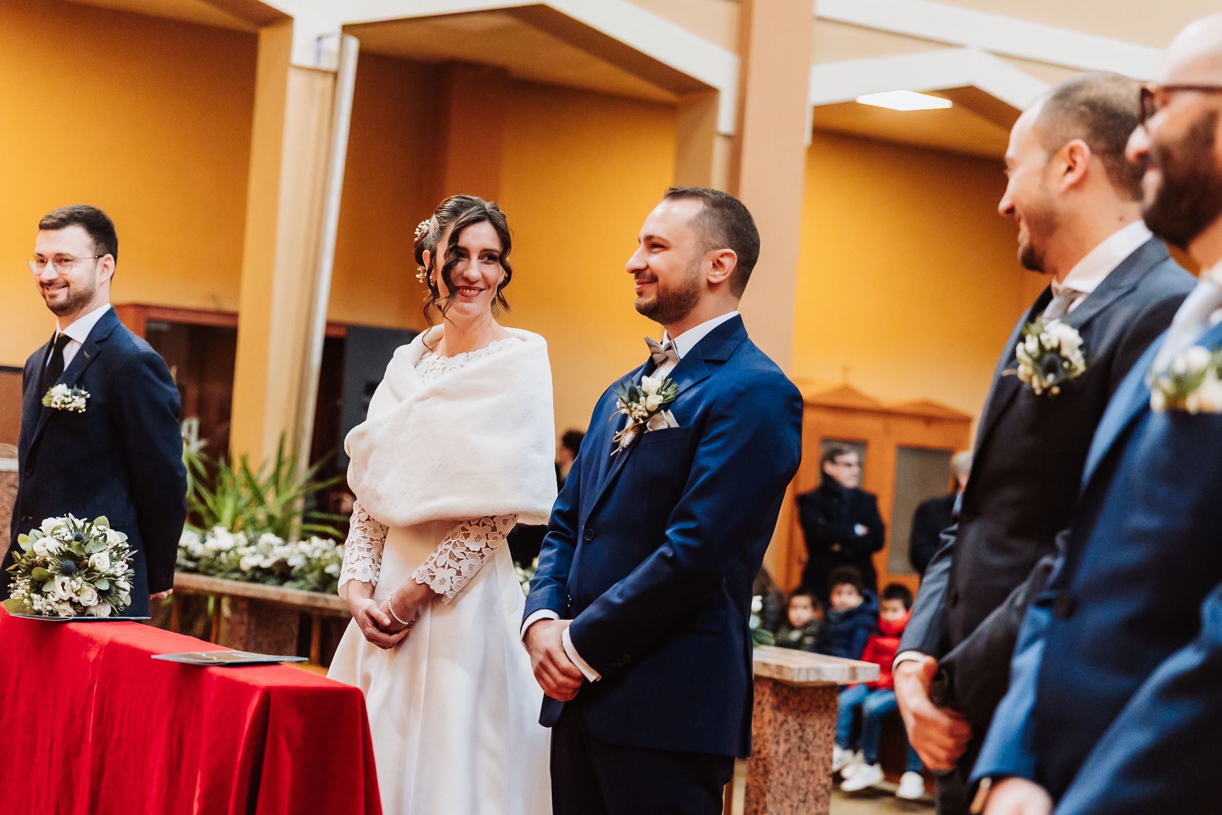 Bride and groom smiling during wedding ceremony, bride in white gown with wrap, groom in navy suit, groomsmen standing nearby.