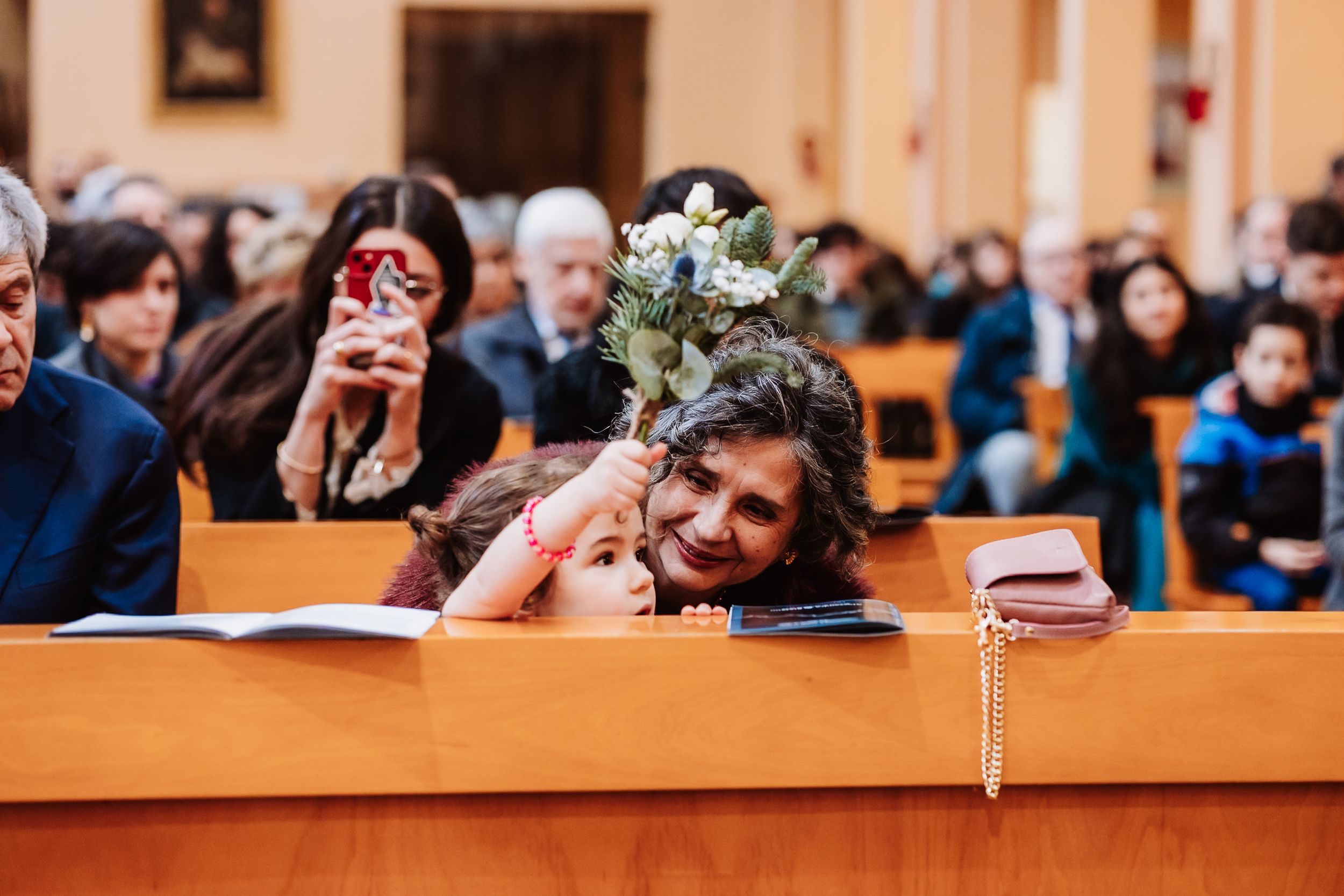 Woman smiling at child holding flowers in church pew during wedding ceremony, guests seated in background.