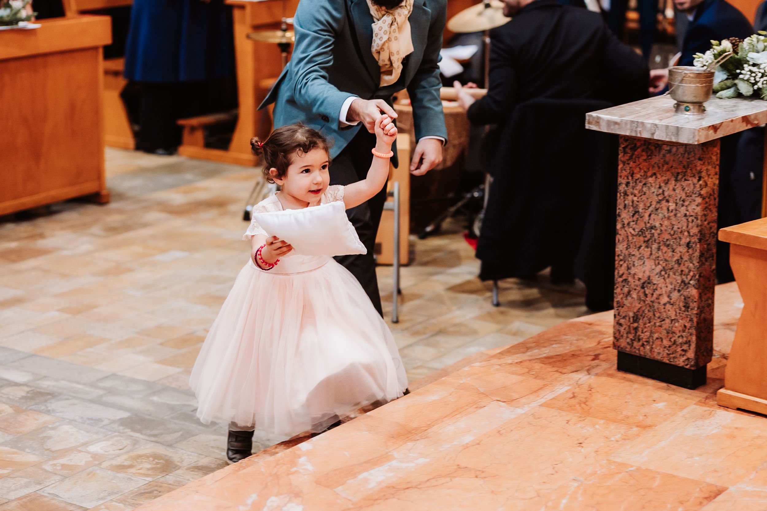 Adorable flower girl in white tulle dress walking down church aisle during wedding ceremony.