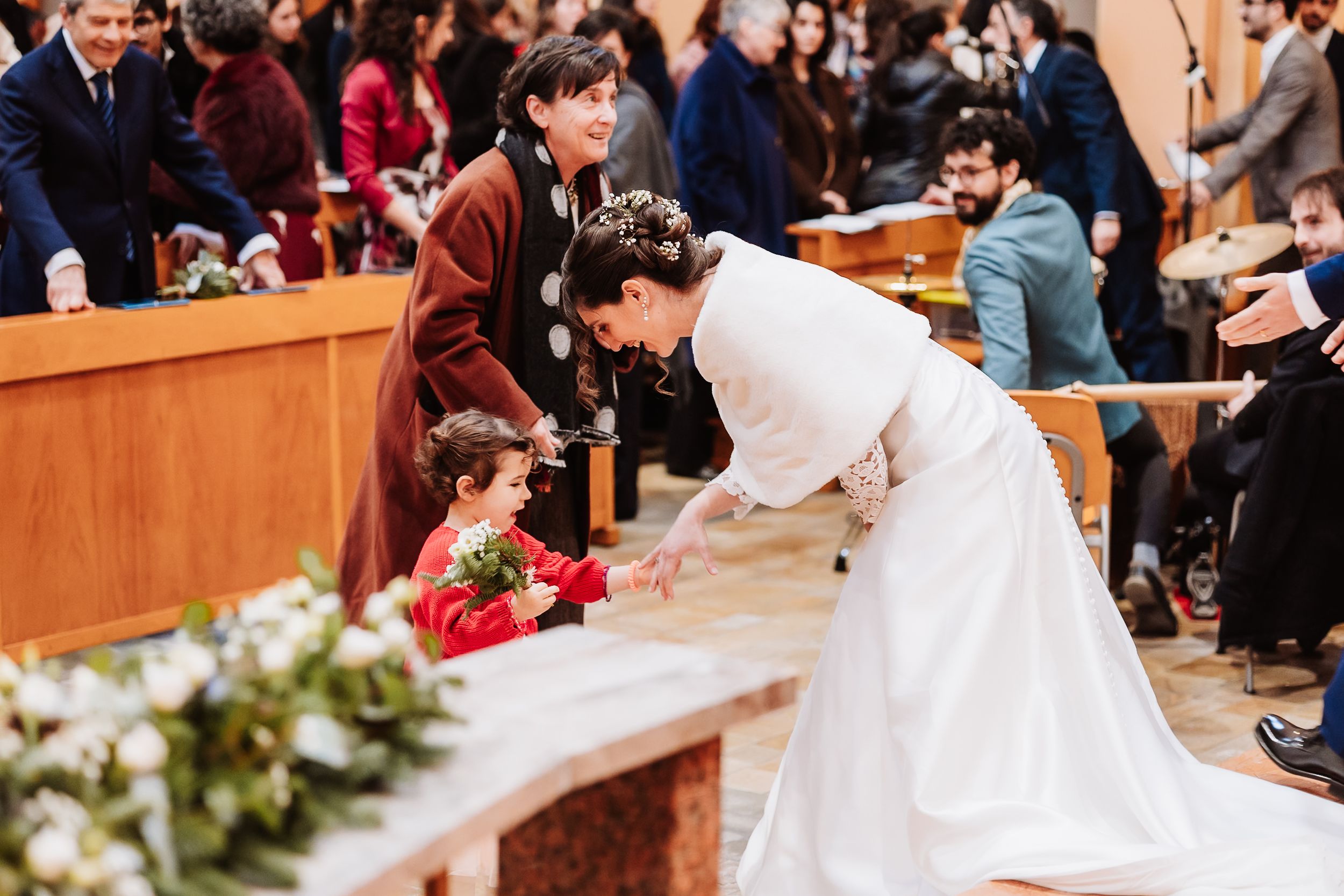 Bride in white gown bends to greet young flower girl in red dress during elegant church wedding ceremony.