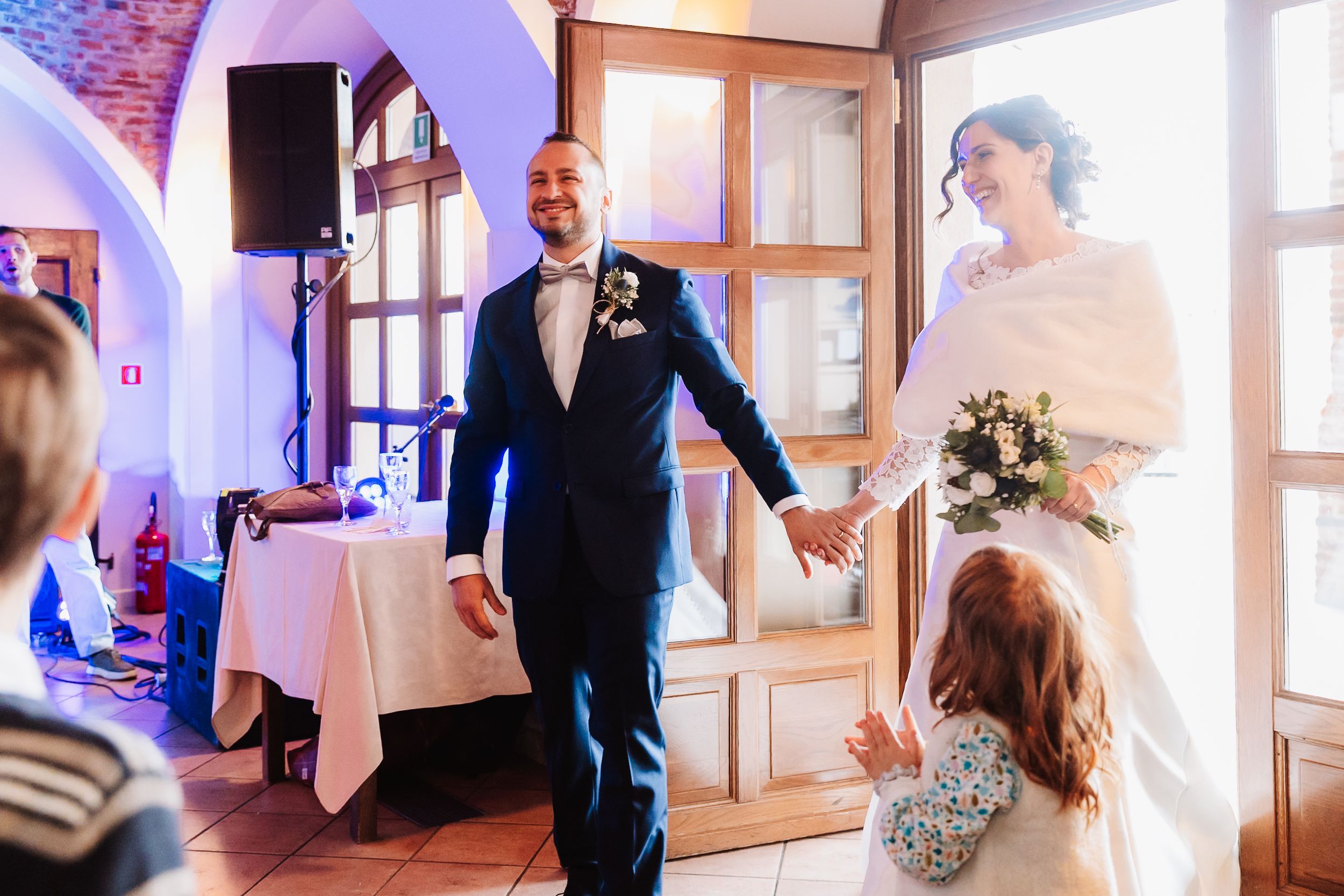 Bride and groom holding hands entering wedding reception hall, bride in white gown with bouquet, groom in navy suit.