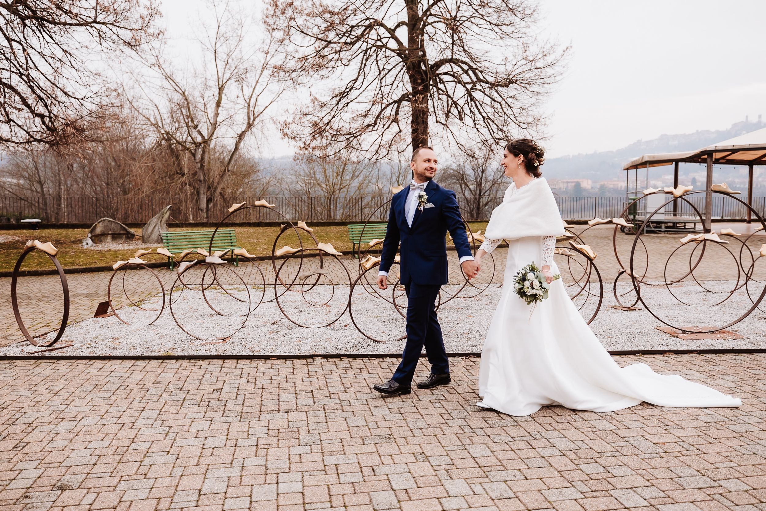 Bride and groom walk hand-in-hand on cobblestone path with decorative metal fence and bare trees in winter wedding photo.
