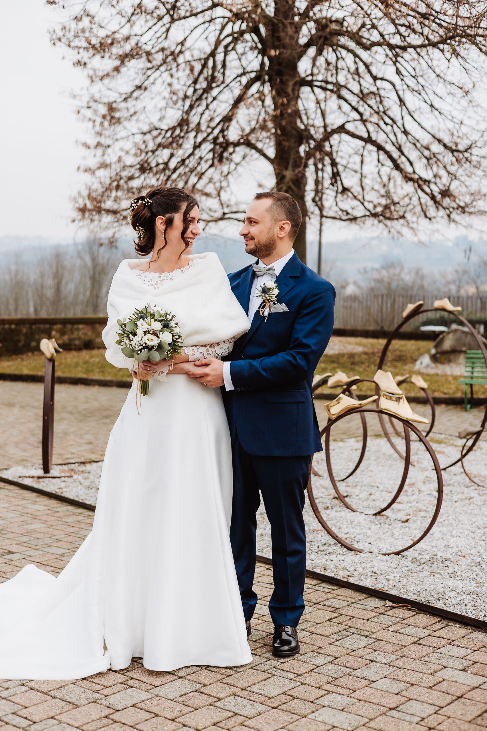 Bride in white gown holding bouquet stands with groom in navy suit outdoors near decorative bicycle sculpture.