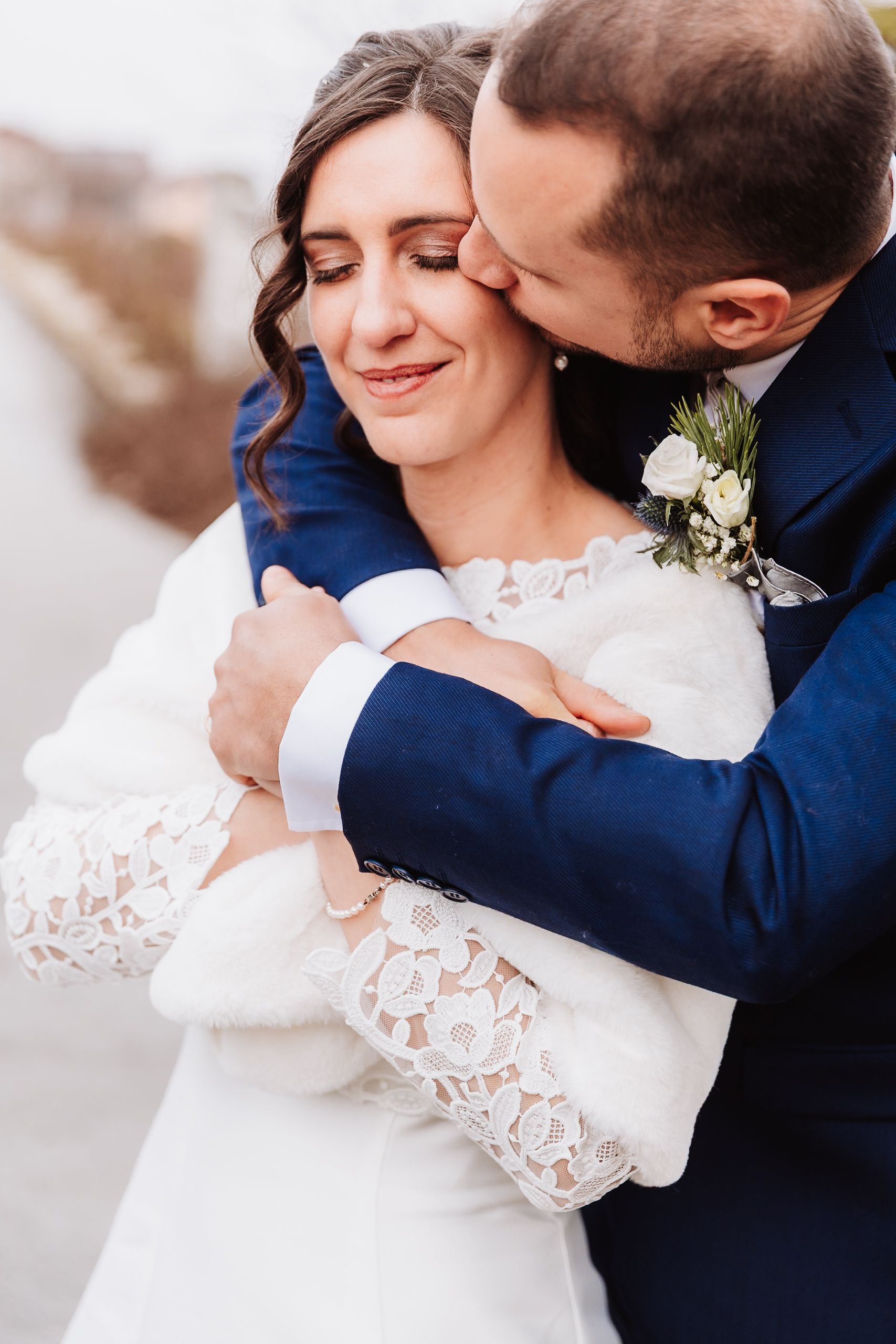 Groom in blue suit embracing bride in lace wedding dress, sharing a tender kiss on her cheek outdoors.