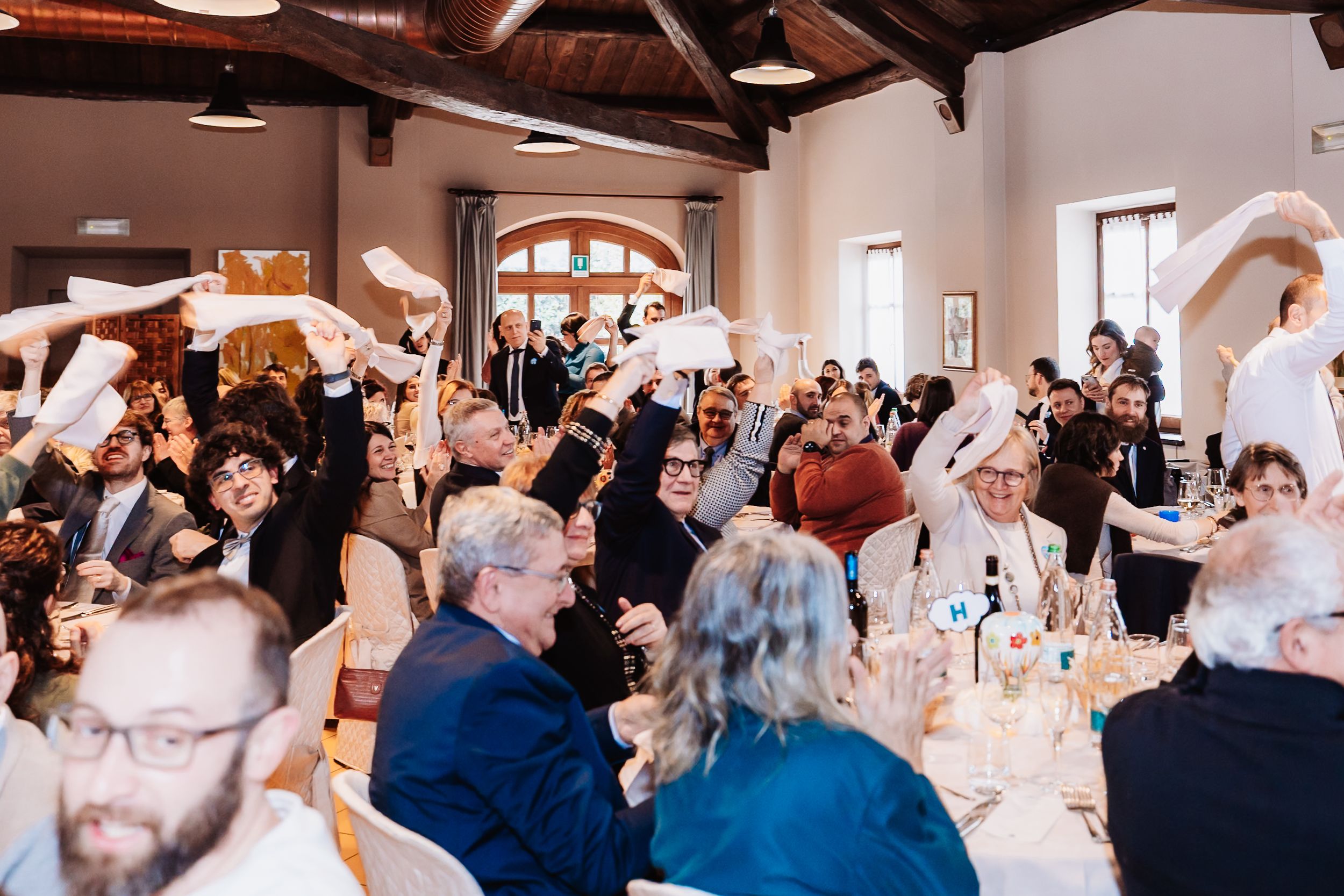 Guests wave white napkins in celebration at an elegant indoor wedding reception dinner in a rustic venue.